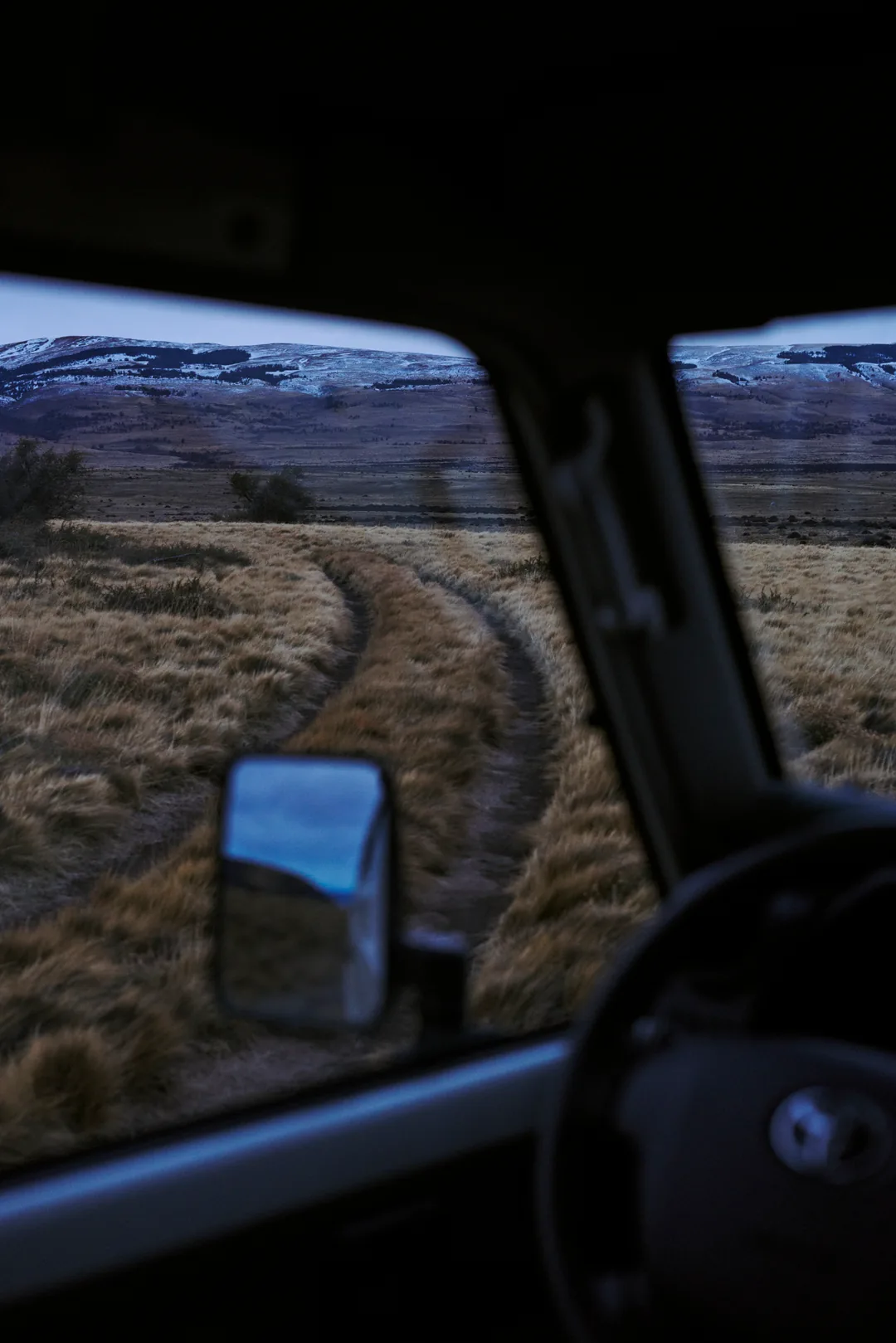 A predawn dirt road toward snow ridges, seen through a truck windshield