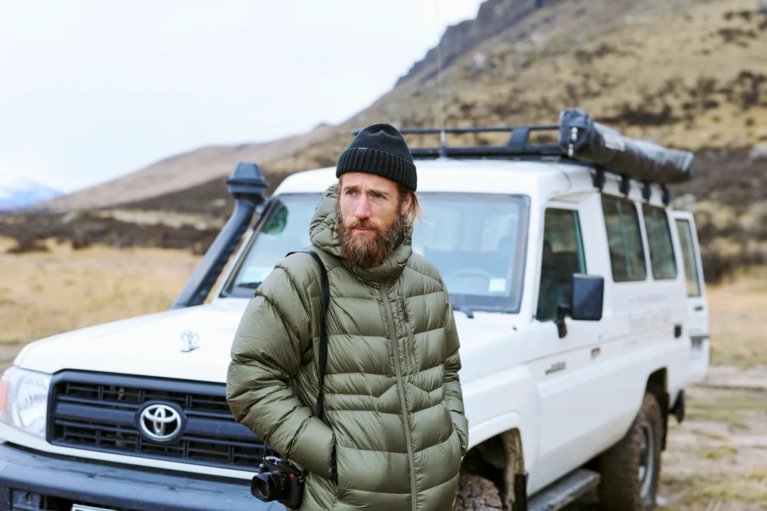 A photographer in an olive puffer and black beanie beside a Toyota troop carrier