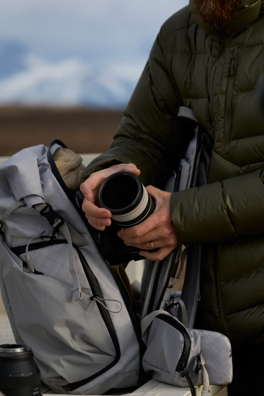 Hands placing a white telephoto lens into a grey backpack, snow mountains behind