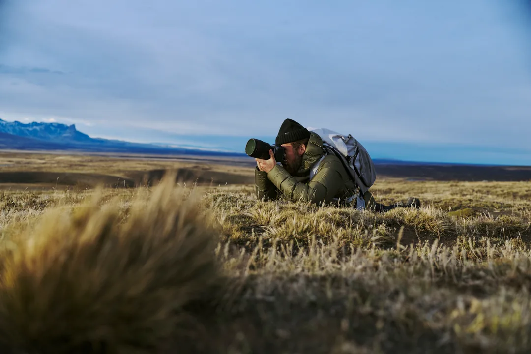 A photographer lying in tundra grass with a telephoto lens, snow ridges behind
