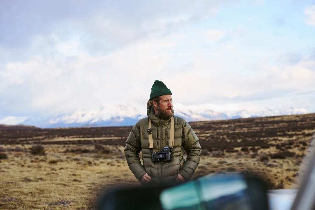 A photographer in a field with a camera on a tan strap, snow mountains far off