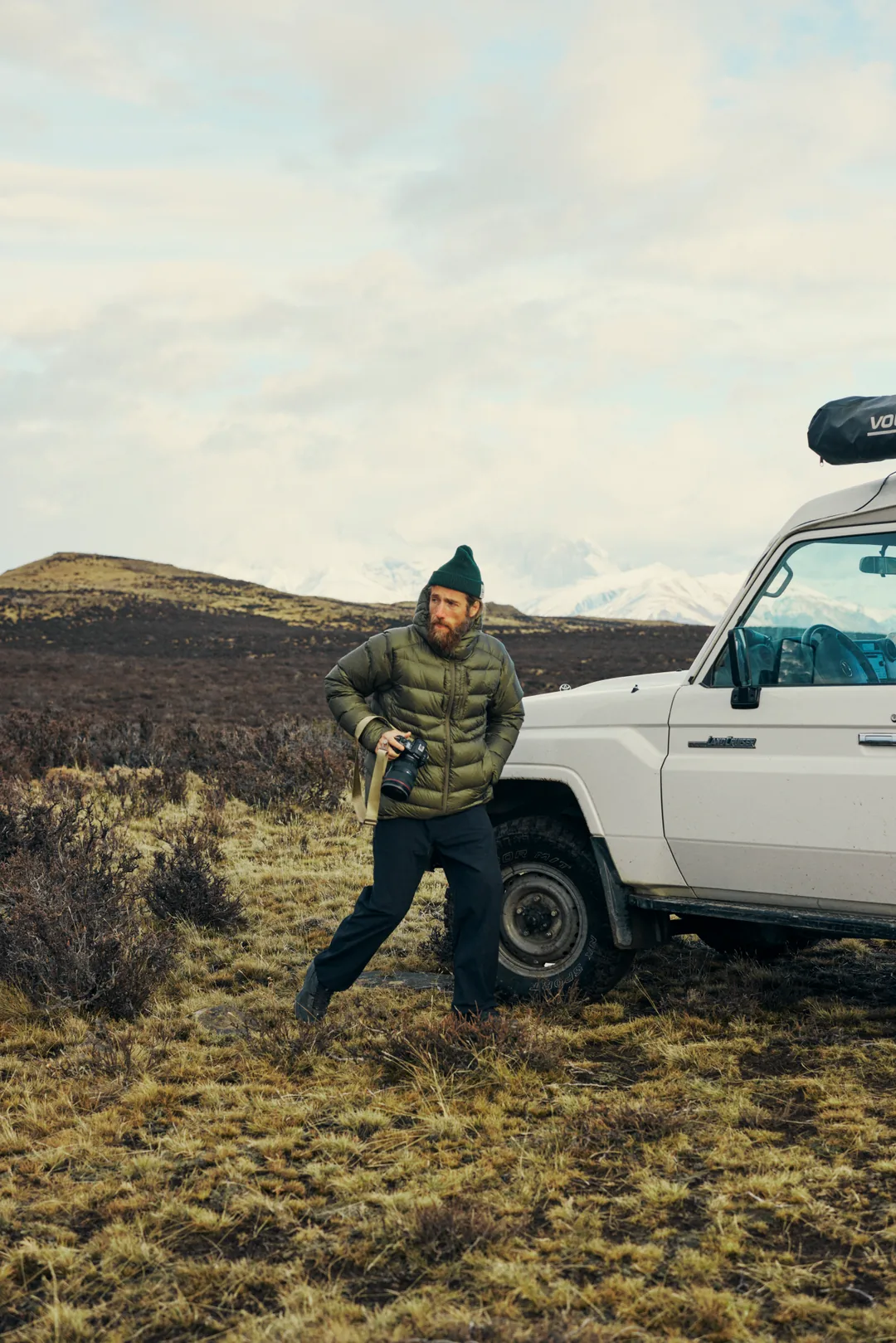 A photographer holding a camera with a white lens beside a Toyota, a snow ridge behind